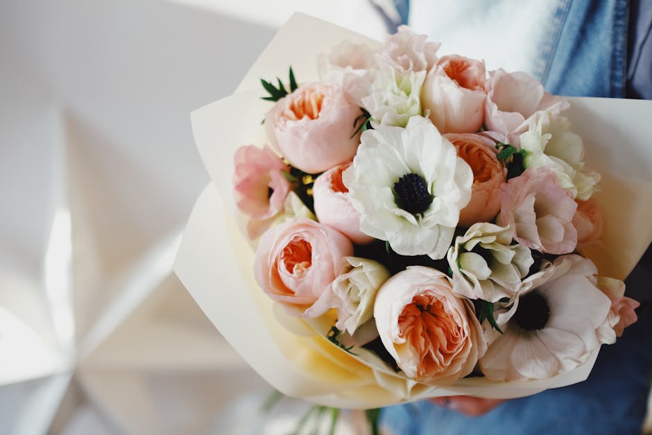 A bouquet of fresh flowers held by a person wearing a blue denim jacket. The arrangement features soft pink roses, white anemones with dark centers, and light pink peonies, all with lush green leaves.