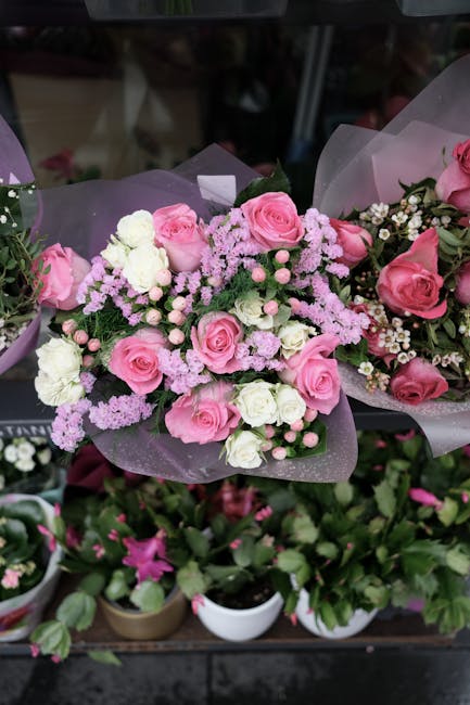 A floral arrangement featuring a bouquet of fresh pink roses, white carnations, and light purple filler flowers, elegantly wrapped in translucent purple floral paper with a satin ribbon. The bouquet i