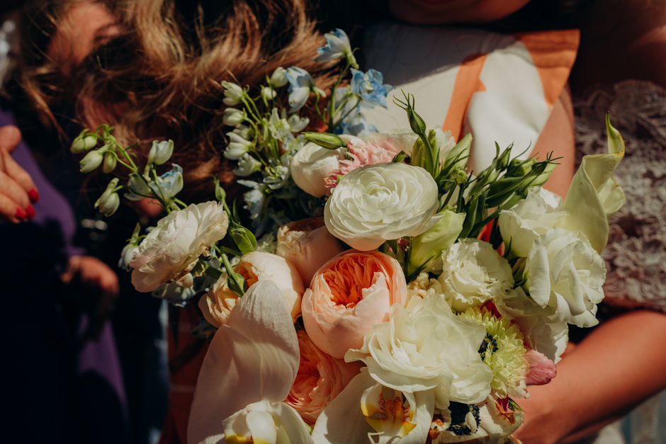 A close-up of a hand-held floral bouquet featuring a variety of fresh flowers, including large white ranunculus, soft peach and cream lisianthus, delicate blue and white delphiniums, and green foliage