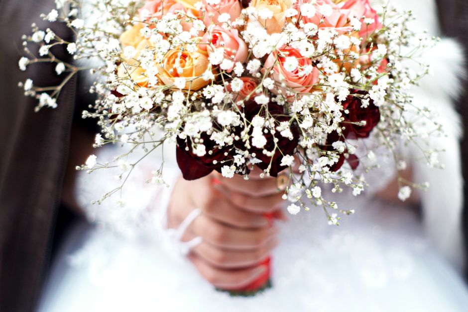 A floral bouquet held by a person, featuring a mix of peach and pink roses, white baby's breath, and deep red flowers, arranged in a rounded, lush style. The bouquet is wrapped in dark red tissue pape