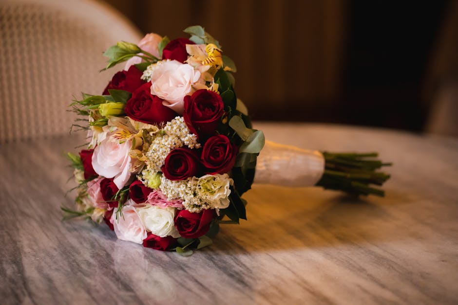 A close-up of a bouquet of fresh flowers arranged with red and pink roses, white and yellow alstroemeria, delicate white spray roses, and sprigs of eucalyptus, wrapped in cream-colored ribbon. The bou