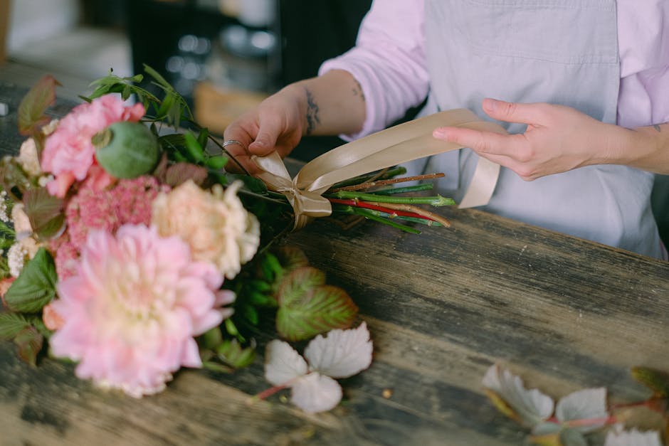 A person with a light pink shirt and tattooed forearm is carefully arranging a bouquet of fresh flowers on a rustic wooden table. The bouquet features large pink peonies, soft peach-colored hydrangeas