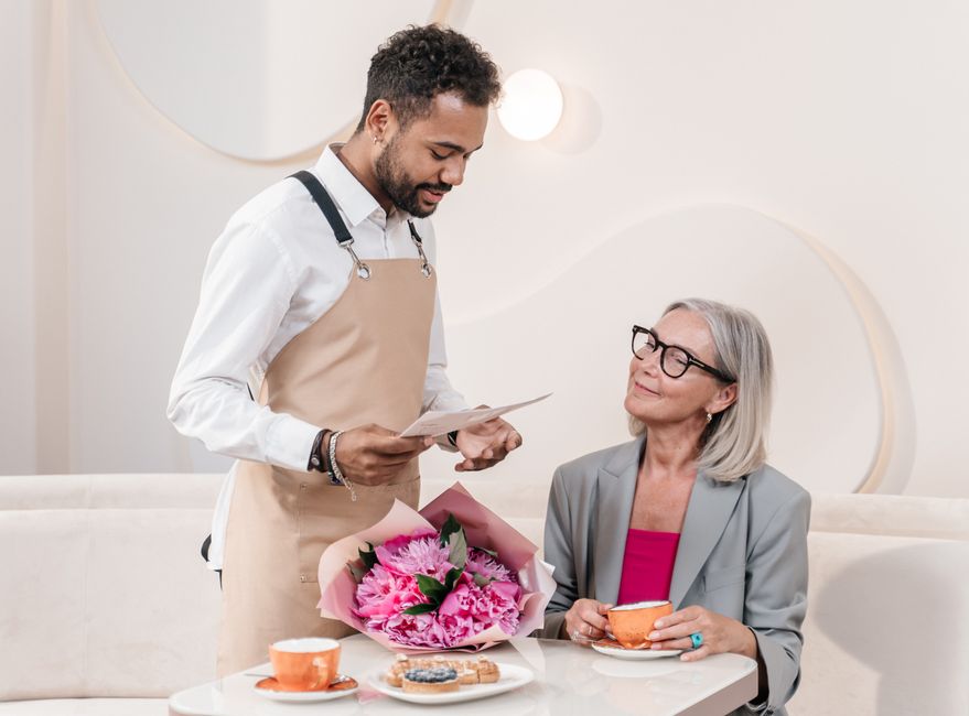 A floral arrangement featuring vibrant pink peonies and carnations wrapped in pink tissue paper, being presented by a male flower delivery courier wearing a beige apron over a white shirt. The arrange