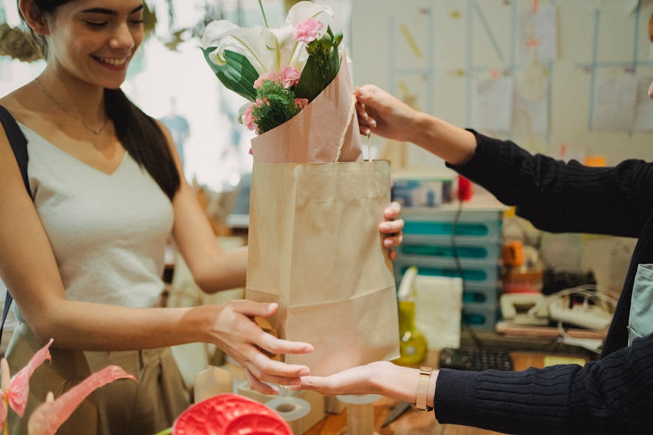 A smiling woman with long dark hair, wearing a light beige sleeveless top, is accepting a flower bouquet from another person dressed in a black sweater in a floral shop. The bouquet is wrapped in soft