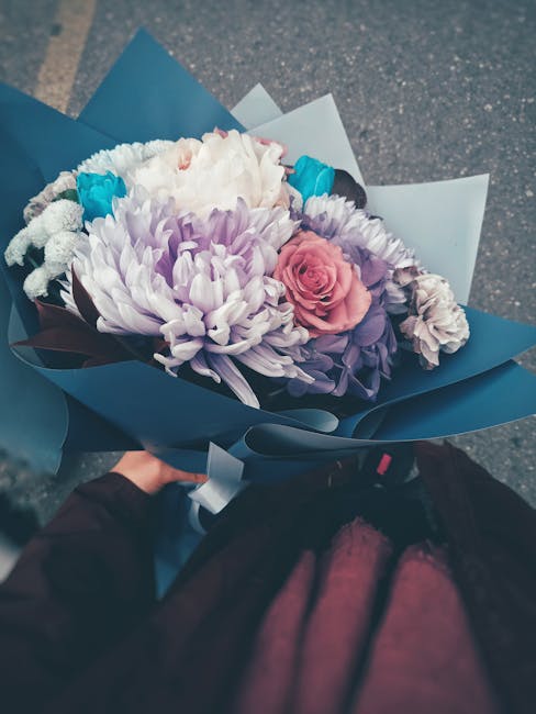 A hand holding a beautifully arranged bouquet of fresh flowers wrapped in dark blue and light blue paper. The bouquet features large, soft white chrysanthemums, delicate pink roses, and light purple l