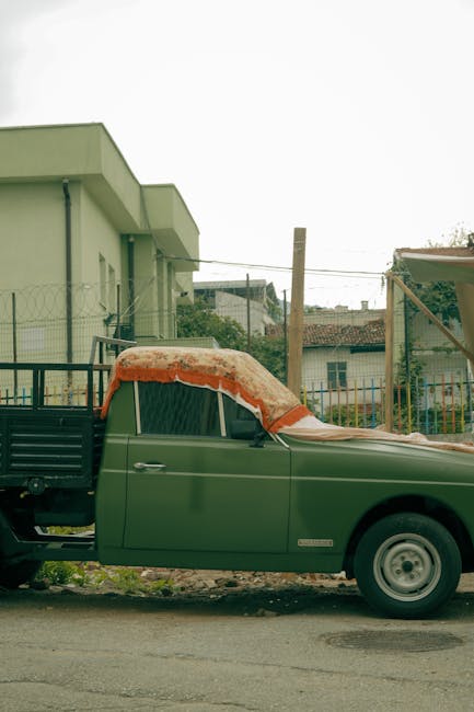 The image depicts a vintage green pickup truck parked outdoors in a residential area. The truck's cab is covered with a brownish-beige tarpaulin, slightly weathered, with some snow on top, suggesting 