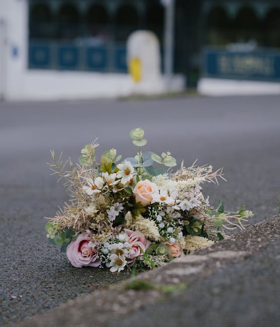 A delicate floral bouquet arranged on the pavement outside, featuring soft pink roses, white daisies, and sprigs of eucalyptus with pale green and cream-colored foliage. The flowers are freshly cut, w
