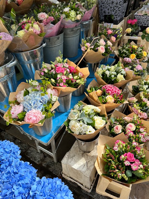 A display of fresh floral arrangements and bouquets at a flower stall, featuring pastel pinks, whites, and light blues. The flowers include roses, hydrangeas, and other seasonal blooms, arranged in sm