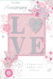In the photograph, an anniversary card rests against a neutral backdrop, its design centred around the timeless sentiment of "LOVE." The letters of the word are raised in a soft emboss, giving them a sculpted appearance that seems to invite touch, like tracing the curve of a flower stem. Each letter is edged in fine glitter, blending blush pink and pearly white tones that glimmer gently rather than flash, much like the subtle sparkle of cut blooms arranged by Flowers Highbury for nearby homes off Blackstock Road. Behind this central word, a blush-pink glow deepens around the letters and then softens into a paler pastel pink that covers the rest of the card, creating a sense of light radiating outward. Surrounding the word, a series of delicate, stylised flowers and curling vines is spread across the surface. The blooms are drawn with fine, graceful lines in shades of light pink, ivory, silver-grey, and touches of metallic silver, echoing the romantic palette of anniversary bouquets inspired by Highbury's garden squares. Some petals and leaves catch the light with a slightly reflective finish, suggesting small areas of foil or extra shimmer. The floral forms weave gently around the central motif, filling the space without ever feeling crowded, suggesting motion and airiness, as though the design could drift off the page. Near the lower portion of the card, an elegant, flowing script carries a tender anniversary message, the cursive strokes echoing the arcs of the vines above. The overall impression is one of calm intimacy and enduring affection, like a quiet evening at home marked by a bouquet on the table and this softly radiant card beside it.